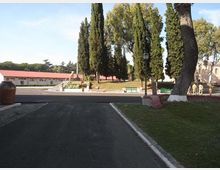 Viale asfaltato che conduce a un’area verde con alti cipressi e grandi alberi, con panchine e un lampione lungo il percorso. Sullo sfondo si vede un edificio basso con tetto rosso e una piccola scalinata in pietra.