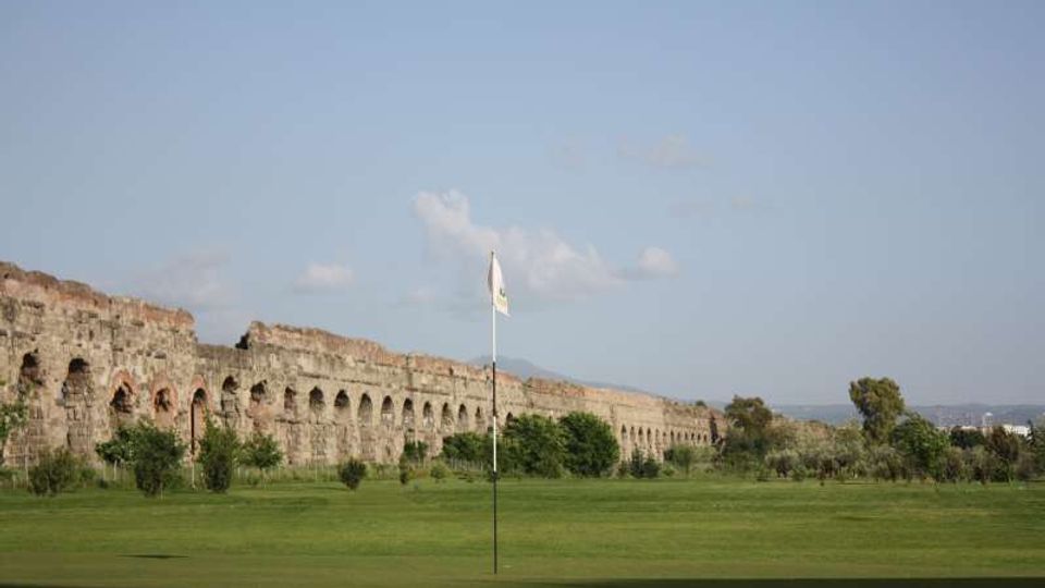 Campo da golf con ampia distesa erbosa e bandierina su asta al centro; sullo sfondo si estende una lunga struttura in pietra con arcate, simile a un acquedotto o muro storico, con alberi sparsi e cielo sereno.