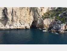 Scogliera costiera di roccia chiara con una piccola grotta alla base, affacciata su mare blu scuro. Sul promontorio sono visibili macchia mediterranea e massi frastagliati con onde che si infrangono vicino alla riva.