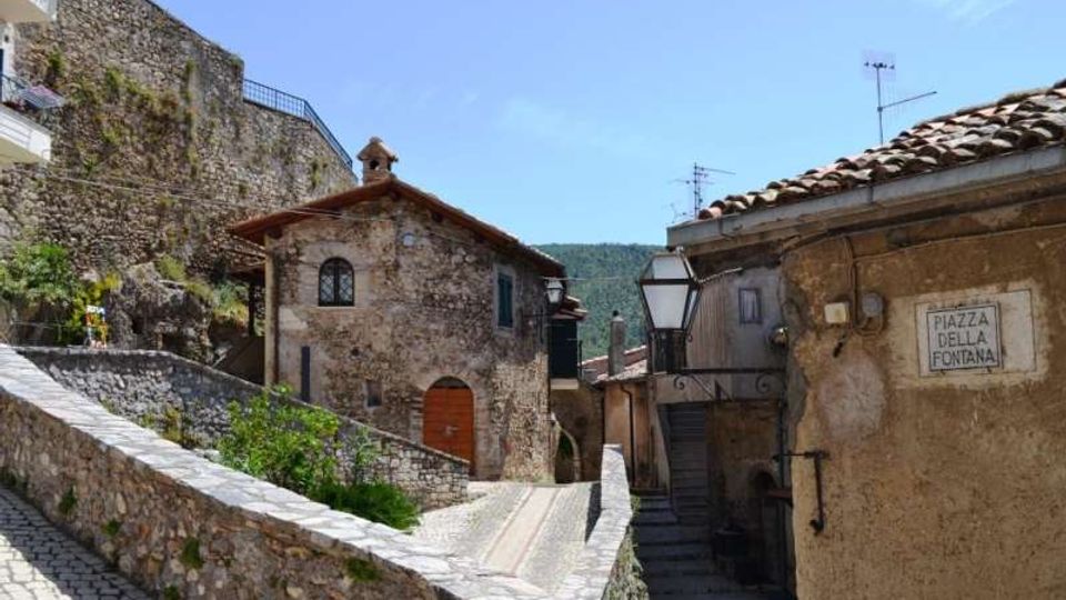 Vicolo in un borgo storico con case in pietra e tetti in coppi, percorso da una scalinata e da una rampa lastricata con muretti laterali. Sulla parete a destra è visibile un’insegna “Piazza della Fontana” e lampioni in ferro, con colline sullo sfondo.
