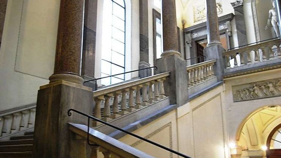 Scalinata interna monumentale in pietra con balaustre, grandi colonne e volte decorate in stile classico. Alte finestre ad arco illuminano un atrio a più livelli con archi e cornici ornamentali.