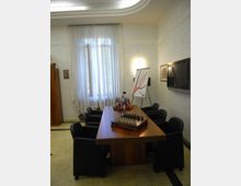 Sala riunioni con lungo tavolo in legno circondato da poltroncine scure, con al centro una scacchiera e piccole bandiere. Sul fondo una grande finestra con tende leggere, mentre sulle pareti ci sono un televisore e un cavalletto con lavagna a fogli mobili.