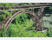 Ponte ad arco in ferro con struttura a traliccio che attraversa una gola, sostenuto da piloni metallici. Intorno si vede una fitta vegetazione e il pendio roccioso della valle.