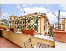 Terrazza su tetto con pavimento in cotto, muretto basso e grigliato verde ad arco con fioriere rettangolari. Sullo sfondo si vedono edifici cittadini in stile classico lungo una strada, con cielo parzialmente nuvoloso.