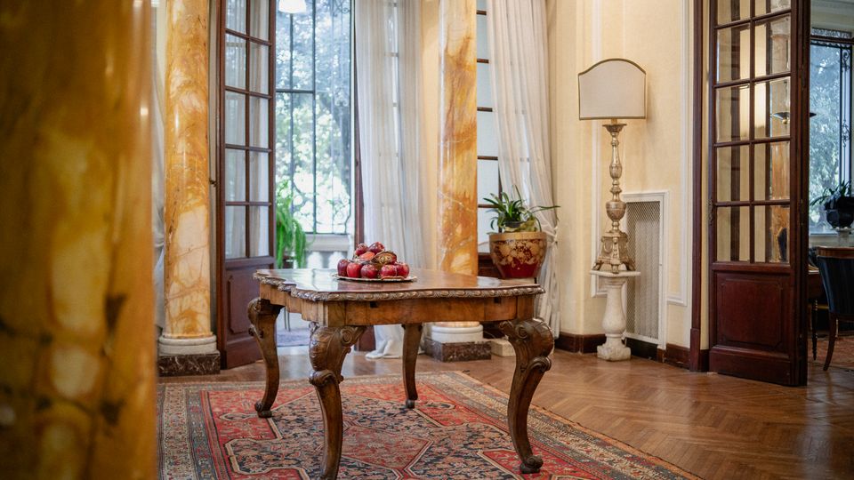 Interno di una villa storica con pavimento in parquet e tappeto persiano, tavolo in legno intagliato al centro e colonne in marmo. Grandi porte e finestre vetrate con tende leggere affacciano sul giardino; a lato si vede una lampada da terra e un vaso con pianta.