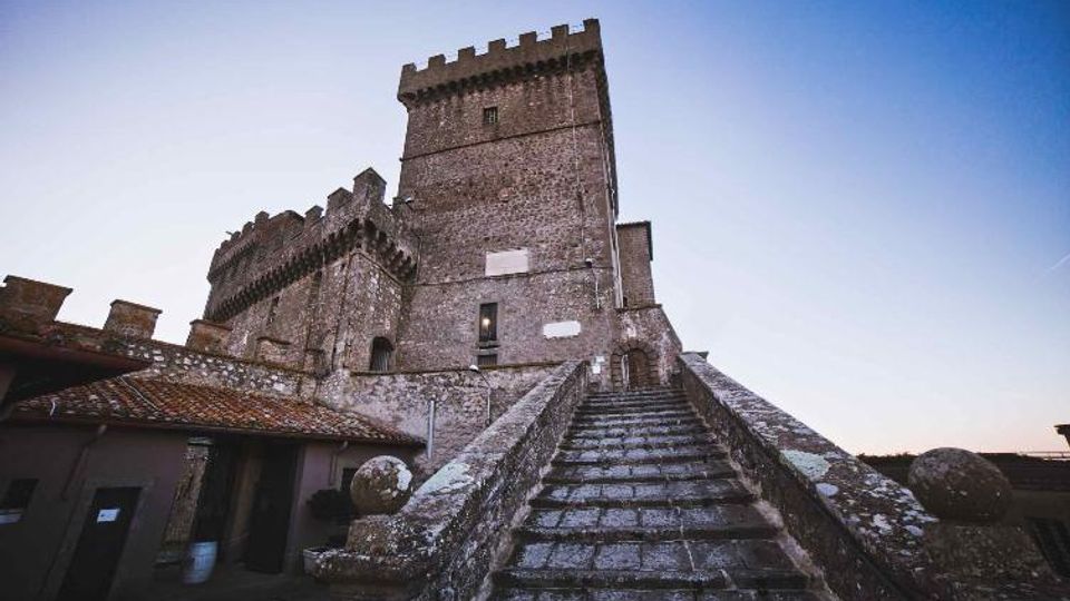 Castello medievale in pietra con torre merlata e mura massicce, visto dal basso. In primo piano una lunga scalinata in pietra con parapetti conduce all’ingresso, con edifici annessi dai tetti in tegole sul lato sinistro.