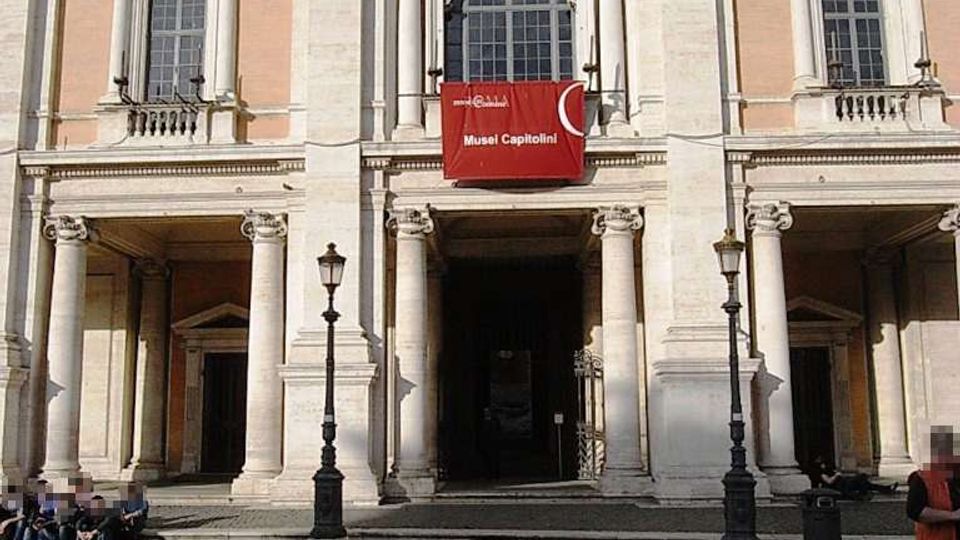 Facciata di un edificio storico in stile classico con portico a colonne e tre ingressi ad arco, grandi finestre incorniciate e decorazioni in pietra chiara su intonaco rosato. Davanti si vede una piazza lastricata con lampioni, e al centro un drappo rosso con la scritta “Musei Capitolini” appeso sotto la finestra.