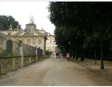 Viale pedonale in un parco, con ampia strada sterrata fiancheggiata da alti alberi e lampioni sul lato destro. A sinistra si vede un edificio storico decorato con facciata in stile classico e recinzione in ferro sopra un muretto.