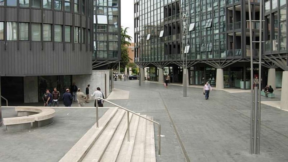 Piazza pedonale moderna pavimentata in pietra, con gradini e corrimano in primo piano e sedute in cemento. Ai lati si affacciano edifici contemporanei con grandi facciate vetrate e strutture metalliche, con lampioni alti lungo il percorso.
