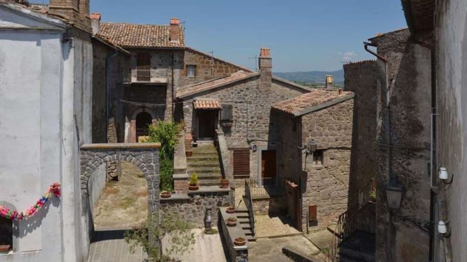 Vista di un piccolo borgo con case in pietra e tetti in tegole, cortili e scalinate in salita tra vicoli stretti. Un arco in muratura e muri intonacati incorniciano la scena, con colline sullo sfondo sotto un cielo sereno.