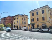 Incrocio urbano con edifici residenziali di tre-quattro piani in tinte pastello, persiane scure e negozi al piano terra con insegne e tende. In primo piano si vedono strisce pedonali, auto parcheggiate e cassonetti lungo il marciapiede sotto un cielo sereno.