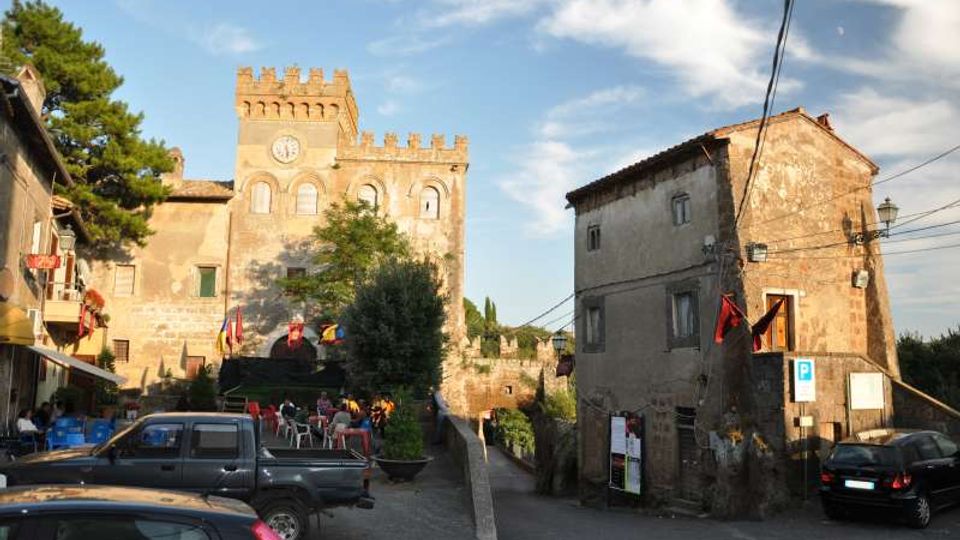 Piazza di un borgo con castello in pietra e torre merlata con orologio sullo sfondo, affiancato da edifici storici intonacati. In primo piano ci sono auto parcheggiate e un passaggio pedonale tra i fabbricati, con alberi e cielo parzialmente nuvoloso.