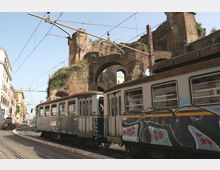 Tram urbano su binari in una strada cittadina, con cavi elettrici aerei e palazzi ai lati. Sullo sfondo si vedono grandi arcate e mura in mattoni di un antico edificio o viadotto, parzialmente coperto da vegetazione.
