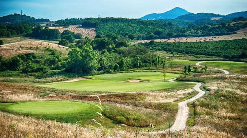 Campo da golf in campagna con fairway verdi ondulati, bunker di sabbia e un sentiero chiaro che serpeggia tra erba alta e cespugli. Sullo sfondo si vedono colline boscose e un rilievo montuoso sotto un cielo azzurro.