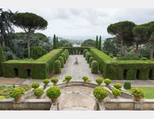 Giardino formale con viali simmetrici delimitati da alte siepi potate e filari di cipressi, con una fontana centrale su pavimentazione in pietra. In primo piano una terrazza con balaustra e vasi di arbusti sferici, circondata da grandi pini e panorama collinare sullo sfondo.