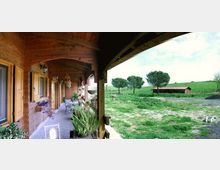 Veranda coperta in legno con porte, finestre e lampade a parete lungo un corridoio con tavolini e piante in vaso. Sul lato aperto si affacciano un prato e una campagna con alberi sparsi e un capannone basso sullo sfondo.