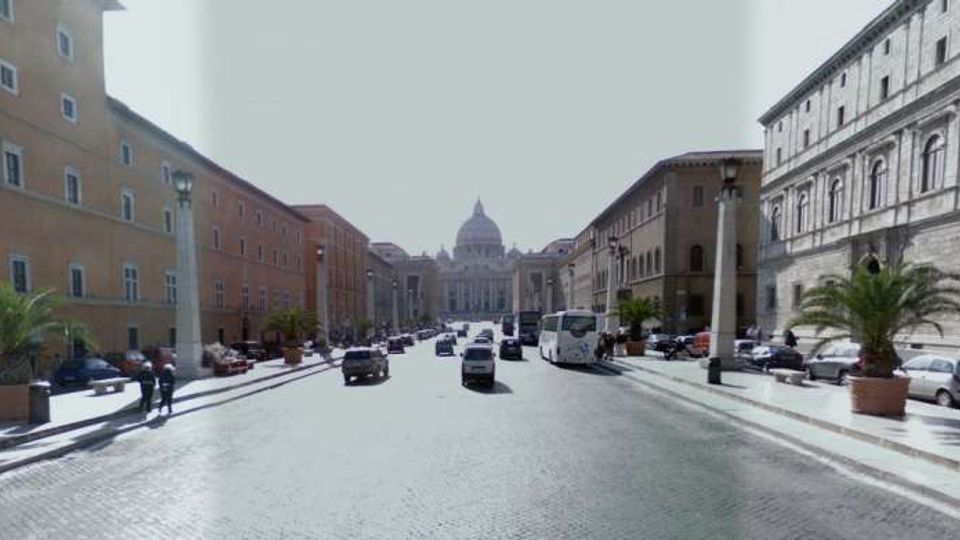 Ampio viale urbano lastricato con traffico e auto parcheggiate ai lati, fiancheggiato da palazzi storici in muratura e lampioni. In fondo alla prospettiva si vede la cupola della Basilica di San Pietro.