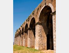 Antico acquedotto in pietra con una serie di grandi archi e pilastri, visto in prospettiva lungo un prato. La struttura in muratura grezza presenta vegetazione che spunta dalla sommità sotto un cielo azzurro.