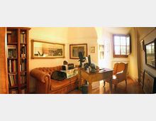 Interno di uno studio domestico con divano in pelle capitonné, libreria alta in legno, scrivania con computer e oggetti decorativi, e una poltrona vicino a una grande finestra. Pareti con quadri incorniciati e pavimento in legno in una stanza illuminata da luce naturale.