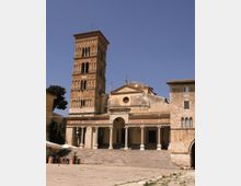 Chiesa storica in pietra con ampia scalinata e portico colonnato, affiancata da un alto campanile in mattoni con arcate su più livelli. Davanti si apre una piazza lastricata, con edifici in pietra ai lati sotto un cielo sereno.