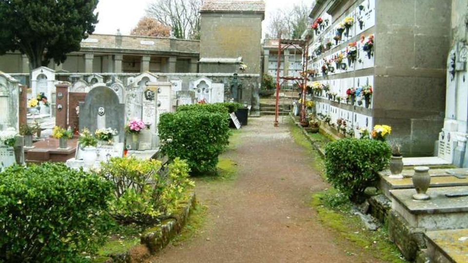 Viale di un cimitero con sentiero in ghiaia, bordato da siepi basse e tombe in pietra con lapidi e fiori. A destra si vede un muro di loculi sovrapposti con decorazioni floreali, con edifici funerari sullo sfondo.
