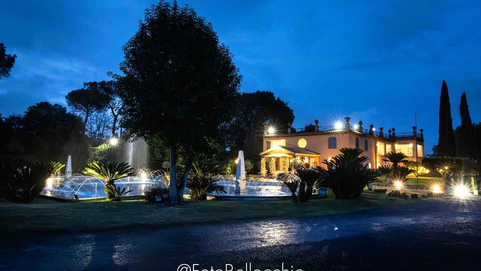 Veduta serale di una villa a due piani illuminata, circondata da un giardino con palme e alberi alti. In primo piano si vede una grande fontana con getti d’acqua e luci, accanto a un vialetto asfaltato bagnato.