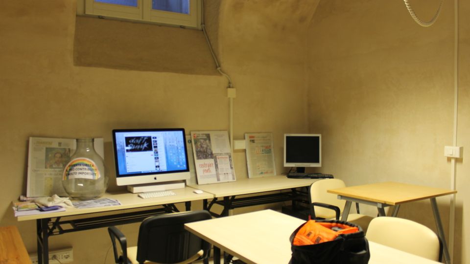 Piccola aula o sala studio con pareti in intonaco e soffitto a volta, illuminata da una finestra alta. Lungo un tavolo sono presenti postazioni con computer e materiali informativi, con banchi e sedie disposti davanti.