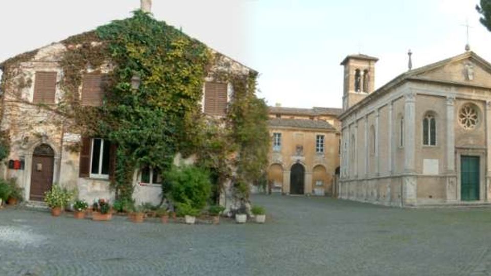 Piazza acciottolata in un borgo storico, circondata da edifici in pietra e intonaco. A destra si vede una chiesa con facciata classica, rosone e campanile, mentre a sinistra c’è una casa rustica coperta di edera con vasi di piante lungo il muro.