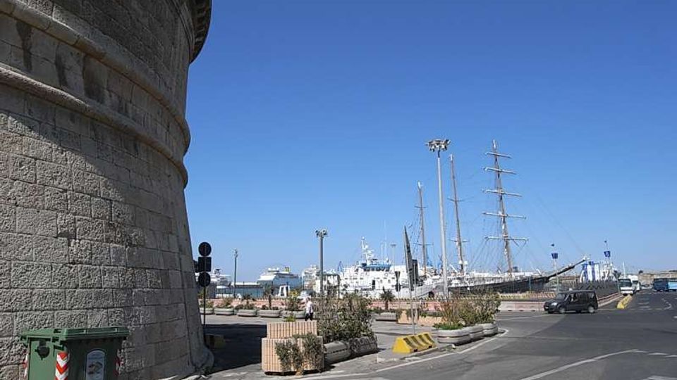 Vista di un’area portuale con strada asfaltata e banchina; a sinistra si vede la parete curva in pietra di una grande struttura circolare. Sullo sfondo sono ormeggiate navi e un grande veliero con alberi alti, con lampioni e fioriere lungo il bordo del porto.