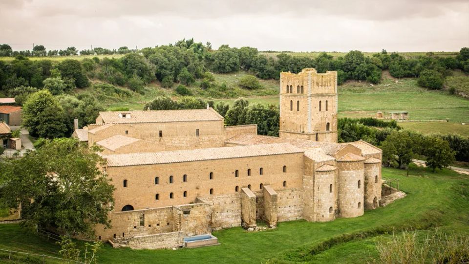 Complesso monastico in pietra con tetti in coppi e una torre campanaria quadrata, circondato da prati e alberi in una zona rurale. Sullo sfondo si vedono colline verdi e un cielo nuvoloso.