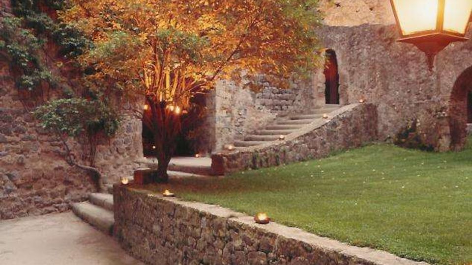 Cortile in pietra di un edificio storico con muri antichi coperti di vegetazione, una scalinata che conduce a un arco e un prato curato con un albero illuminato. In primo piano si vede un vialetto con muretto in pietra e un lampione in stile classico.