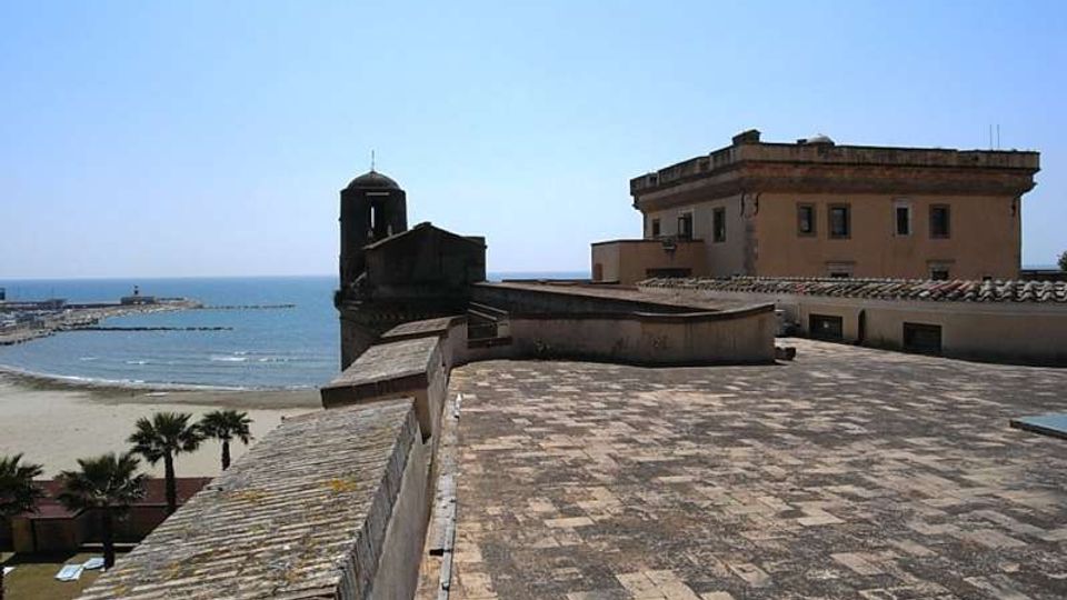 Terrazza in pietra su una fortificazione costiera, con muretto di cinta e edificio storico con torre campanaria; sullo sfondo si vedono la spiaggia e il mare. A destra compare un edificio rettangolare intonacato con tetto basso e finestre affacciate verso la costa.