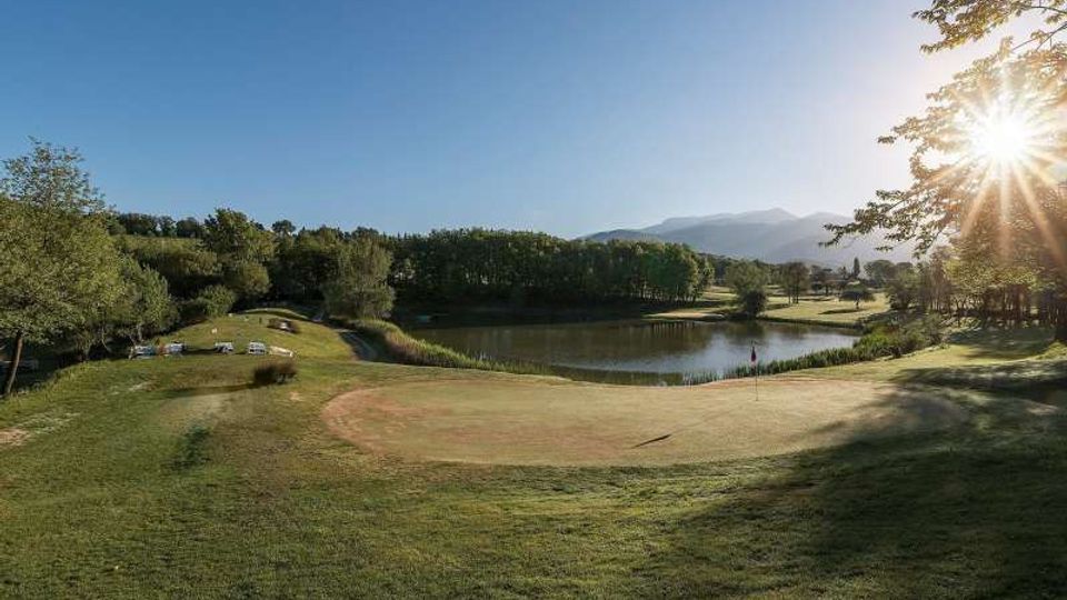 Campo da golf con green in primo piano e bandierina, affacciato su un laghetto circondato da alberi e prati. Sullo sfondo si vedono colline e montagne sotto un cielo sereno con il sole basso all’orizzonte.
