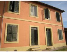 Facciata di una casa a due piani intonacata color salmone con cornici beige, persiane verdi e due porte d’ingresso al piano terra con piccoli gradini. Tetto a falde con sporto e finestre disposte regolarmente lungo la parete esterna.