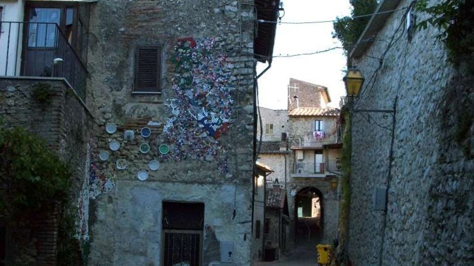 Vicolo stretto in un borgo storico, con pavimentazione in pietra e alti muri di case in muratura grezza. In fondo si vede un arco che attraversa la strada, mentre su una facciata a sinistra sono presenti decorazioni a mosaico e piatti ceramici.
