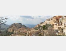 Panorama di un borgo collinare con case in pietra dai tetti in tegole, addossate lungo un crinale e su pendii terrazzati. Sullo sfondo si vedono colline e vallate, con rami spogli in primo piano e un gruppo di edifici più grandi in cima al colle.