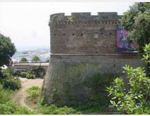Torre cilindrica di una fortificazione in mattoni con merlature e base inclinata in pietra, circondata da vegetazione. Sullo sfondo si intravede la costa e un’area portuale, con un grande cartellone sul lato destro della torre.