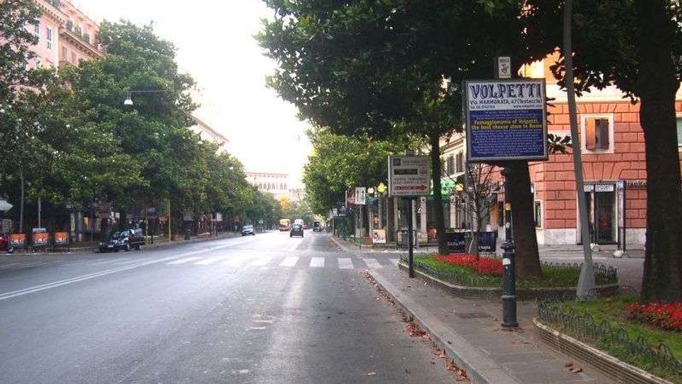Ampio viale urbano alberato con carreggiata a più corsie e attraversamento pedonale, affiancato da palazzi cittadini e marciapiedi. In primo piano si vedono cartelli stradali e un’aiuola con fiori rossi lungo il bordo della strada.