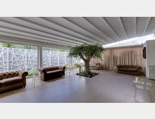 Ampio lounge interno con soffitto a travi bianche e grandi vetrate su tre lati, affacciate su un patio con pannelli geometrici. Al centro c’è un albero in una fioriera, con divani Chesterfield in pelle e piccoli tavolini metallici disposti lungo le pareti e davanti a tende chiare.