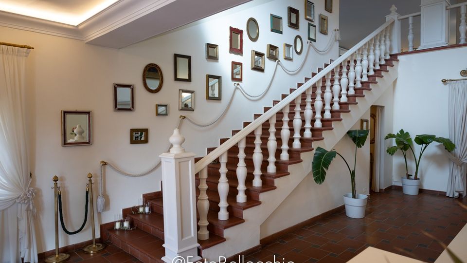 Interno di una hall con grande scala in legno e ringhiera bianca, parete decorata con specchi e cornici di varie dimensioni lungo la salita. Pavimento in cotto, tende bianche alle finestre e piante in vaso ai lati della scala.