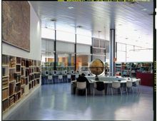 Interno di una biblioteca moderna con scaffali di libri lungo le pareti, postazioni con computer e tavoli di studio. Ampie vetrate e soffitto a pannelli illuminano lo spazio, con un grande globo su un tavolo centrale.