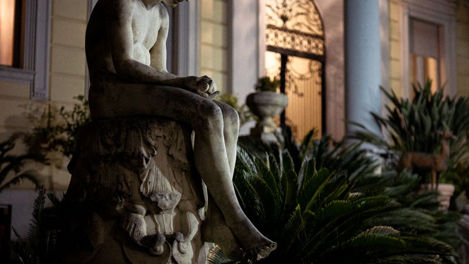 Statua in pietra su piedistallo in un giardino con palme nane e vegetazione, davanti alla facciata di una villa con colonne e finestre. Sullo sfondo si vede un portale ad arco con cancello decorato, illuminato in luce notturna.