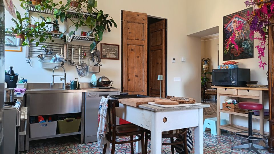 Cucina abitata con pavimento in piastrelle decorative e isola centrale in legno bianco con piano in marmo e sgabelli. A sinistra ci sono lavello e piano in acciaio con mensole piene di piante e utensili appesi, mentre a destra un carrello con microonde e una porta in legno conduce a un altro ambiente.