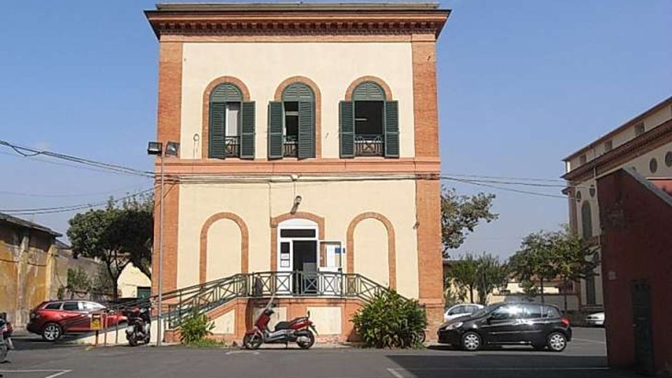 Edificio pubblico a due piani in stile tradizionale, con facciata color crema e cornici in mattoni, finestre ad arco con persiane verdi e ingresso centrale raggiungibile da una rampa con ringhiera. Davanti si apre un piazzale asfaltato con parcheggi e cavi aerei, circondato da alcuni alberi e costruzioni laterali.