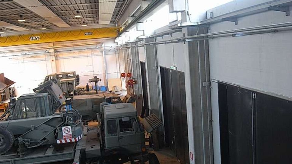 Interno di un grande capannone industriale tipo officina, con soffitto alto in metallo e lucernari laterali che illuminano l’ambiente. Lungo le pareti ci sono ampie porte scorrevoli e all’interno sono presenti vari veicoli e macchinari pesanti parcheggiati sul pavimento in cemento.