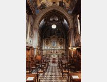 Interno di una cappella o piccola chiesa con navata unica, pavimento a scacchi e file di sedie in legno rivolte verso l’altare decorato. Volta a botte con affreschi e archi ornamentali, pareti con pannellature lignee e dipinti, e rosone circolare sopra il presbiterio.