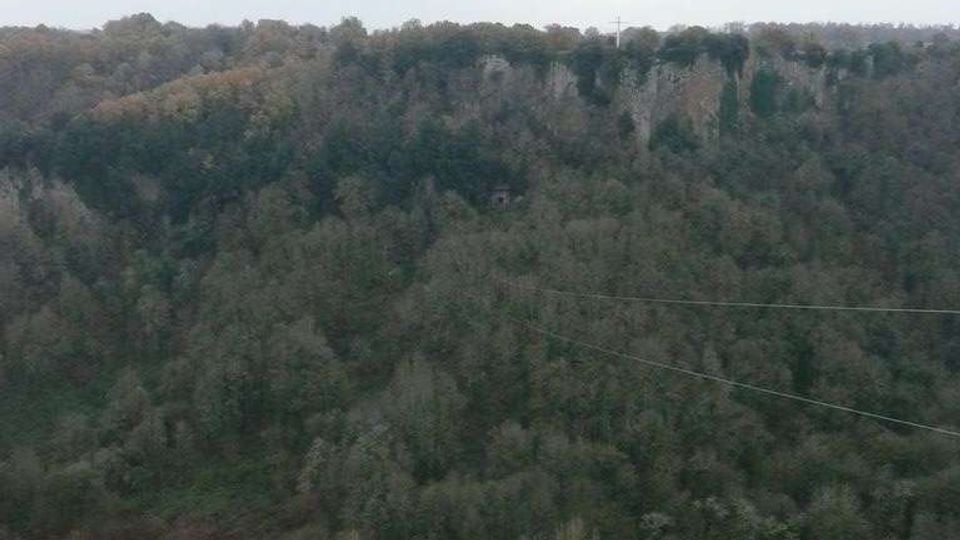 Veduta panoramica di una valle boscosa con fitta vegetazione e una scarpata rocciosa lungo la cresta in lontananza. Il cielo è coperto e alcune linee elettriche attraversano la scena.