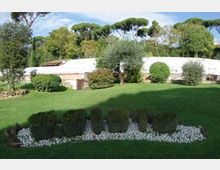 Giardino curato con ampio prato verde, siepi potate e aiuole bordate di ciottoli bianchi in primo piano. Sullo sfondo si vede una lunga struttura bassa con copertura bianca, circondata da alberi e pini marittimi.
