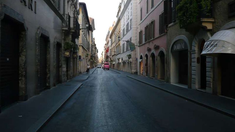 Strada cittadina stretta fiancheggiata da edifici storici a più piani con facciate in intonaco, persiane e portici ad arco con ingressi di negozi. Asfalto scuro con marciapiedi ai lati e vista prospettica verso un incrocio in lontananza.