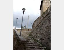 Scalinata in pietra che sale tra alti muri di contenimento in muratura grezza, affiancata da lampioni lungo il percorso. In cima si intravede un edificio con facciata intonacata e tetto spiovente sotto un cielo nuvoloso.